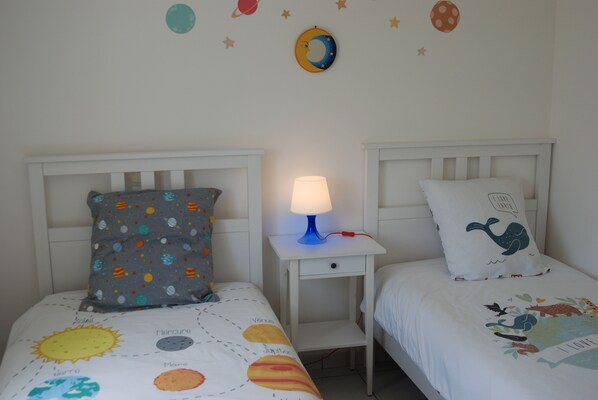 A child's bedroom with two beds, each with a unique pillow design, a nightstand with a lamp, and a wall decorated with space-themed decals.