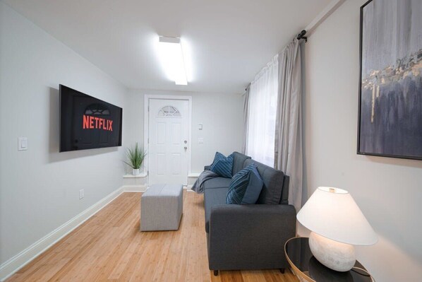 Un salon avec un canapé gris, une lampe blanche et un téléviseur fixé au mur qui affiche le logo de Netflix.