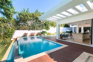 Un espace piscine extérieur moderne, doté d’une terrasse en bois, d’un espace salon couvert et d’un bar.