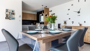 Et moderne spiseområde med et træbord dækket til fire personer, omgivet af sorte stole. Et stort vægur er synligt, og en potteplante tilføjer et strejf af grønt.