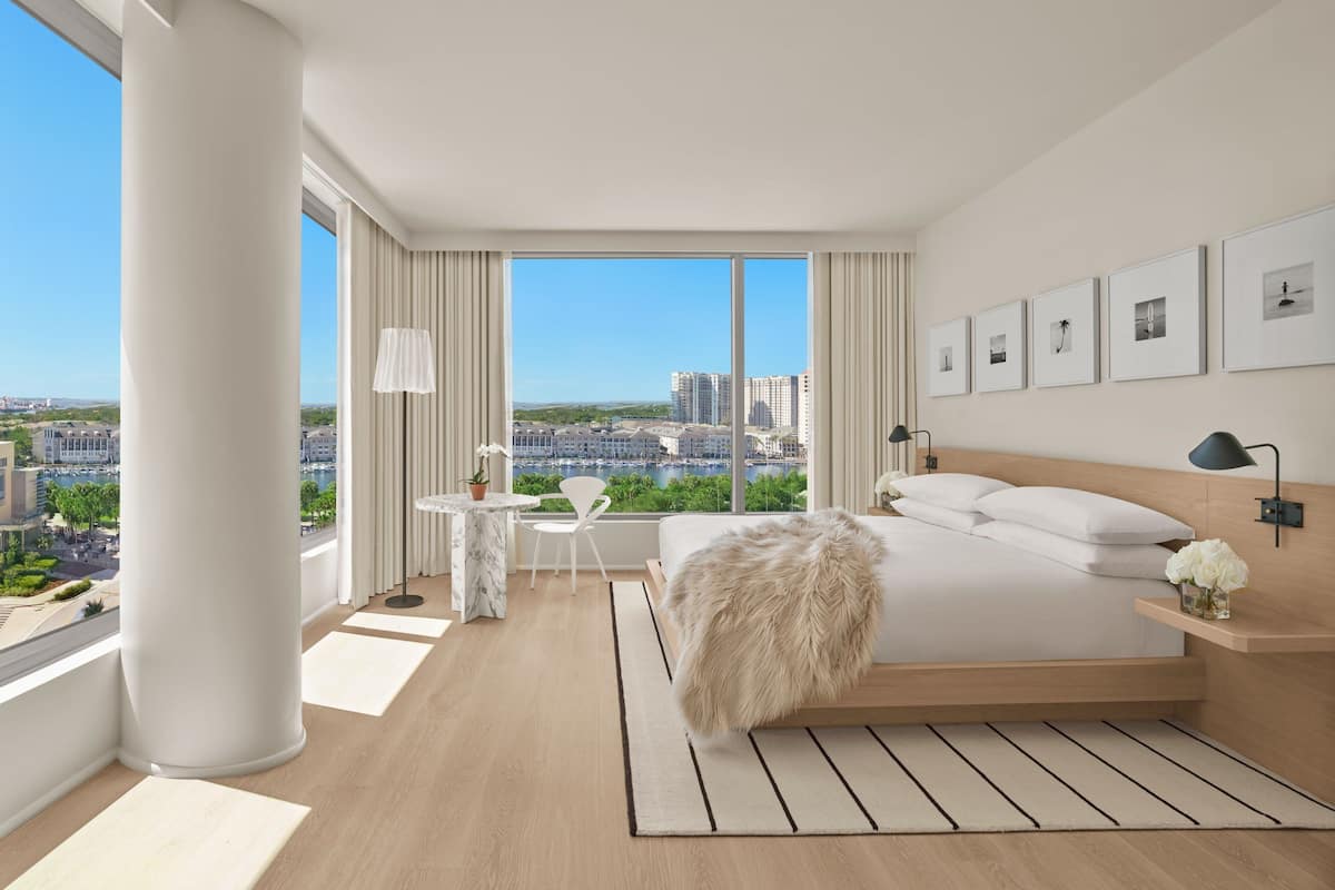 A modern hotel room with a large bed, a desk, a chair, and a view of the city.
