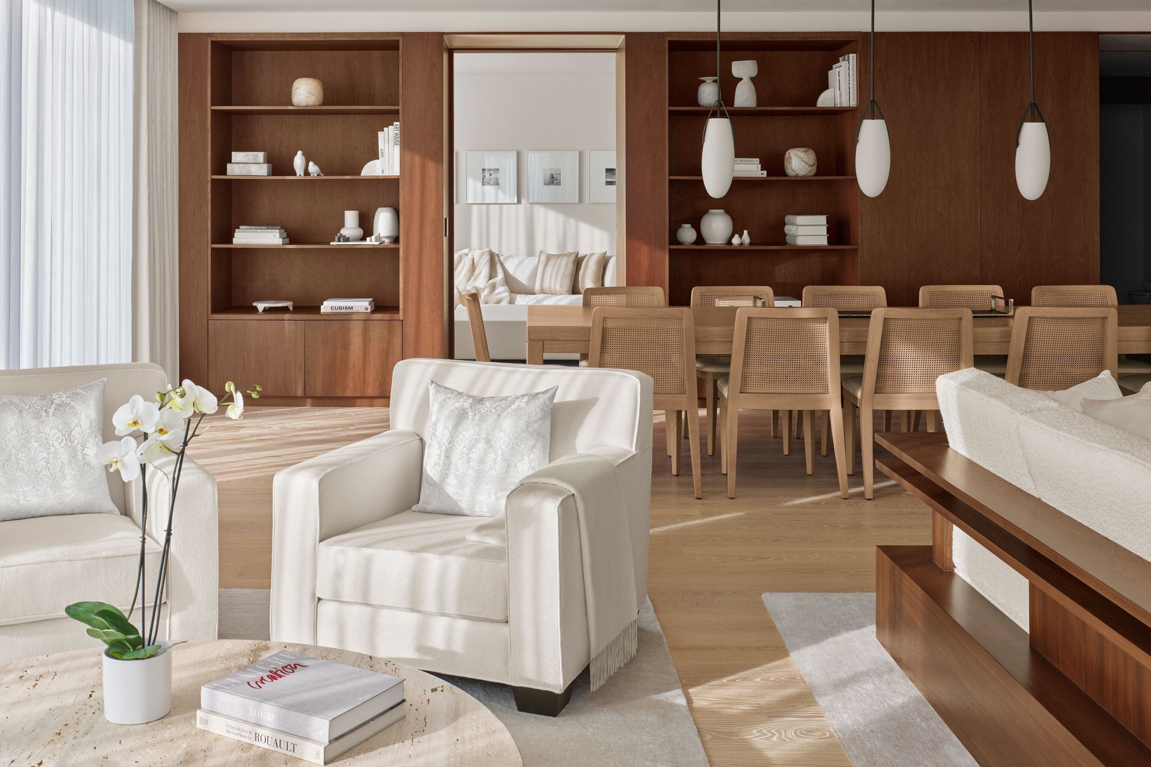 A modern living room with a sofa, armchairs, a coffee table, and a dining area with chairs and a table. There are bookshelves with decorative items and a large window with curtains.