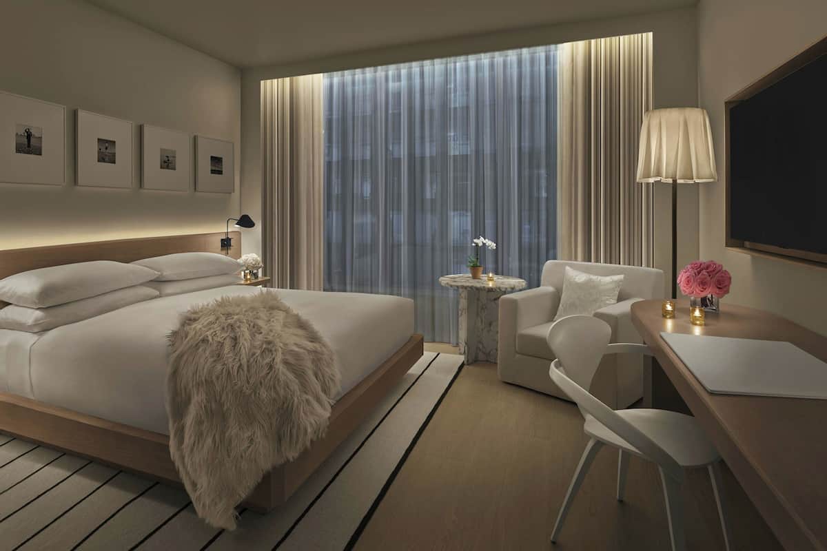 A modern hotel room with a large bed, a desk with a chair, a small round table with a lamp, and a large window with curtains.