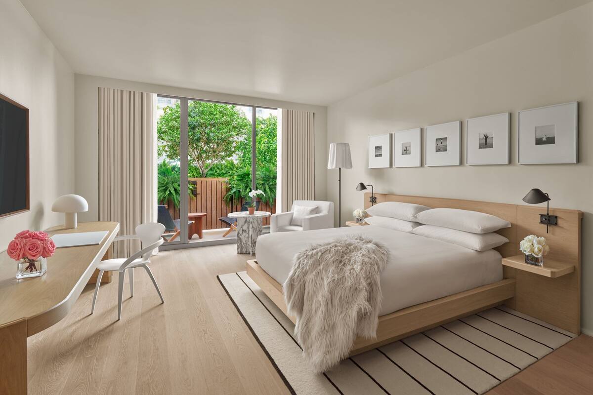 A modern hotel room with a large bed, a desk with a chair, a small table with a chair, and a view of greenery outside.