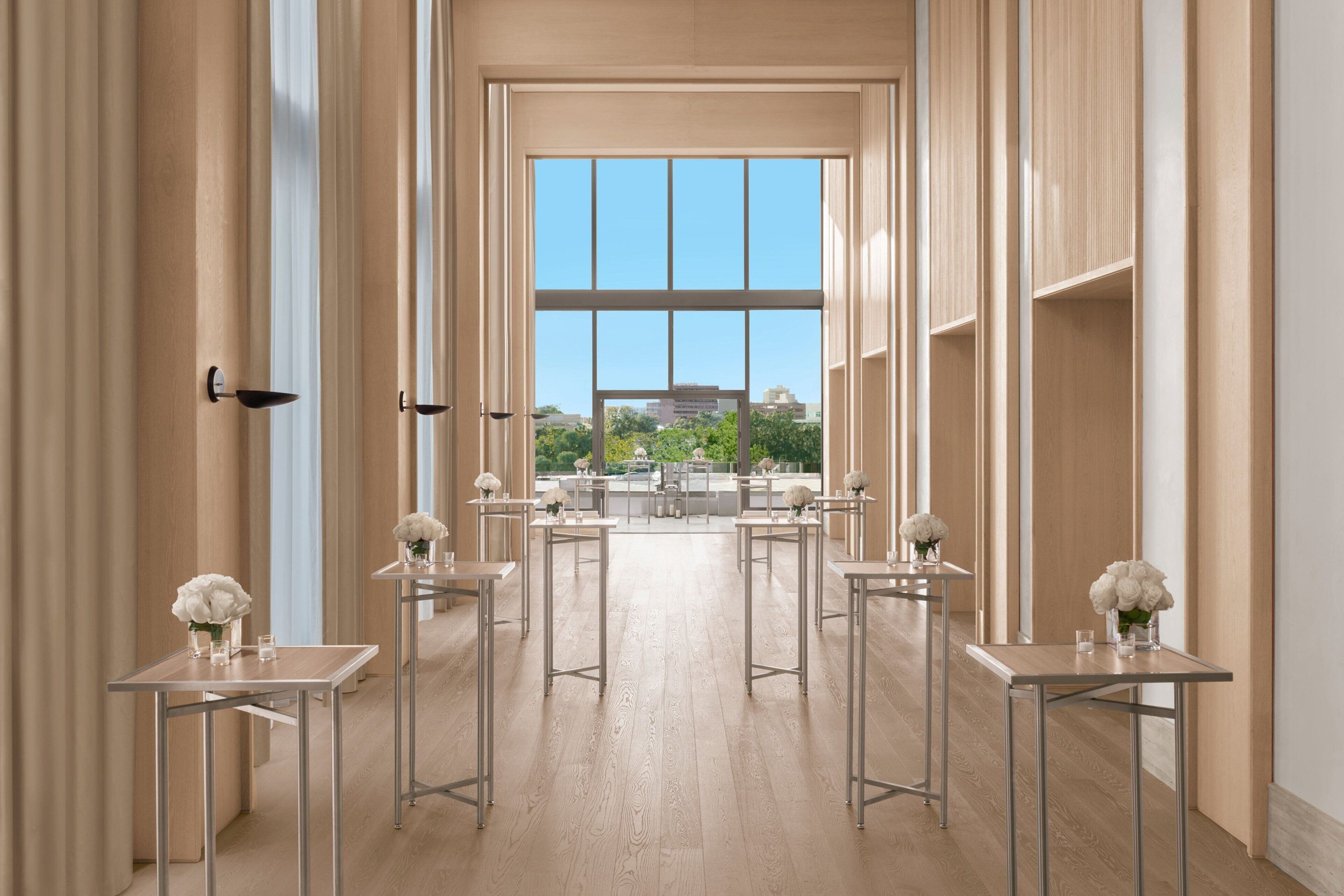 A spacious hotel corridor with a clear view of the outdoors, featuring multiple tables with vases and a modern design.