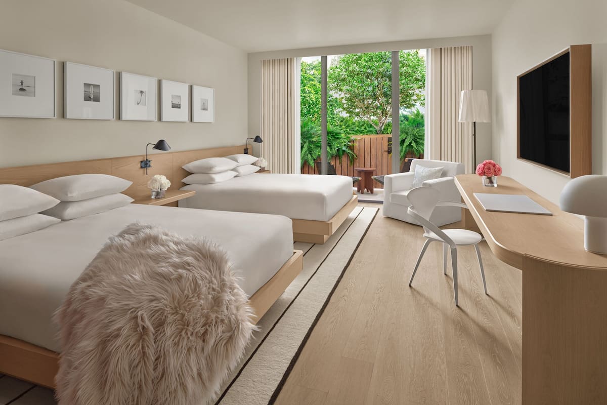 A modern hotel room with two beds, a desk, a chair, and a large window with a view of greenery.