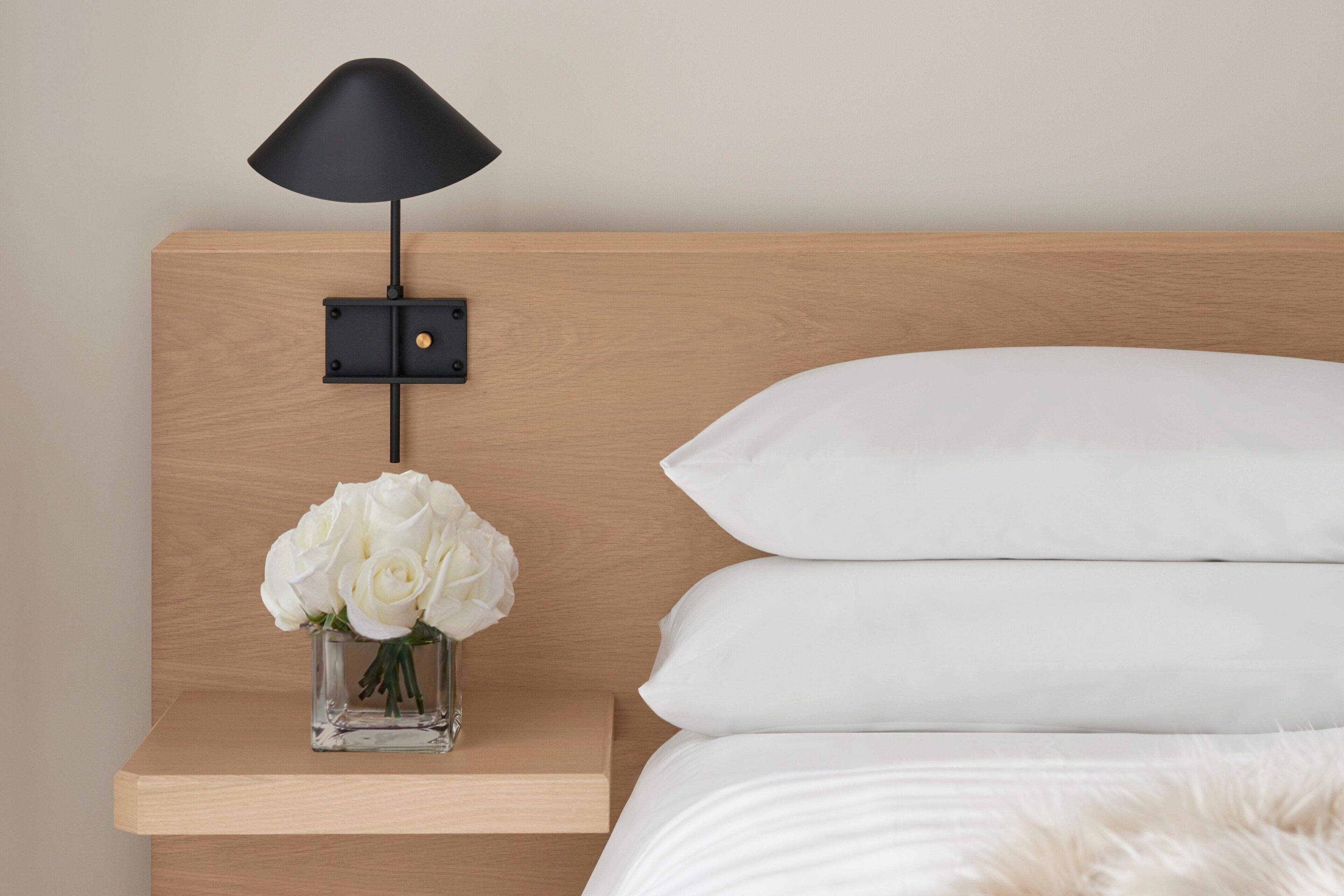 A neatly made bed with white bedding, a small bedside table with a black lamp, and a vase of white flowers.