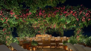 Un área de comedor al aire libre con mesas y sillas, rodeada de exuberante follaje y plantas en flor.