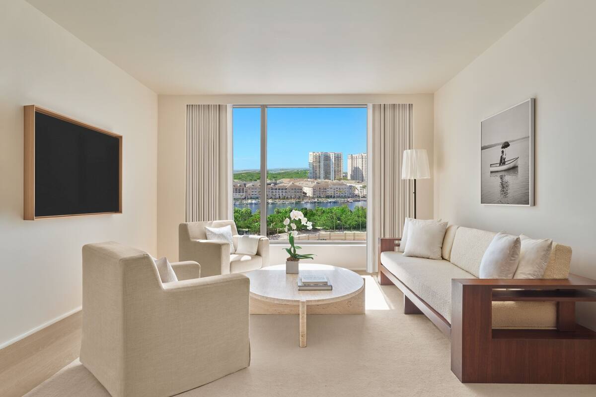 A modern hotel room with a large window, a round coffee table, a sofa, and a TV mounted on the wall.