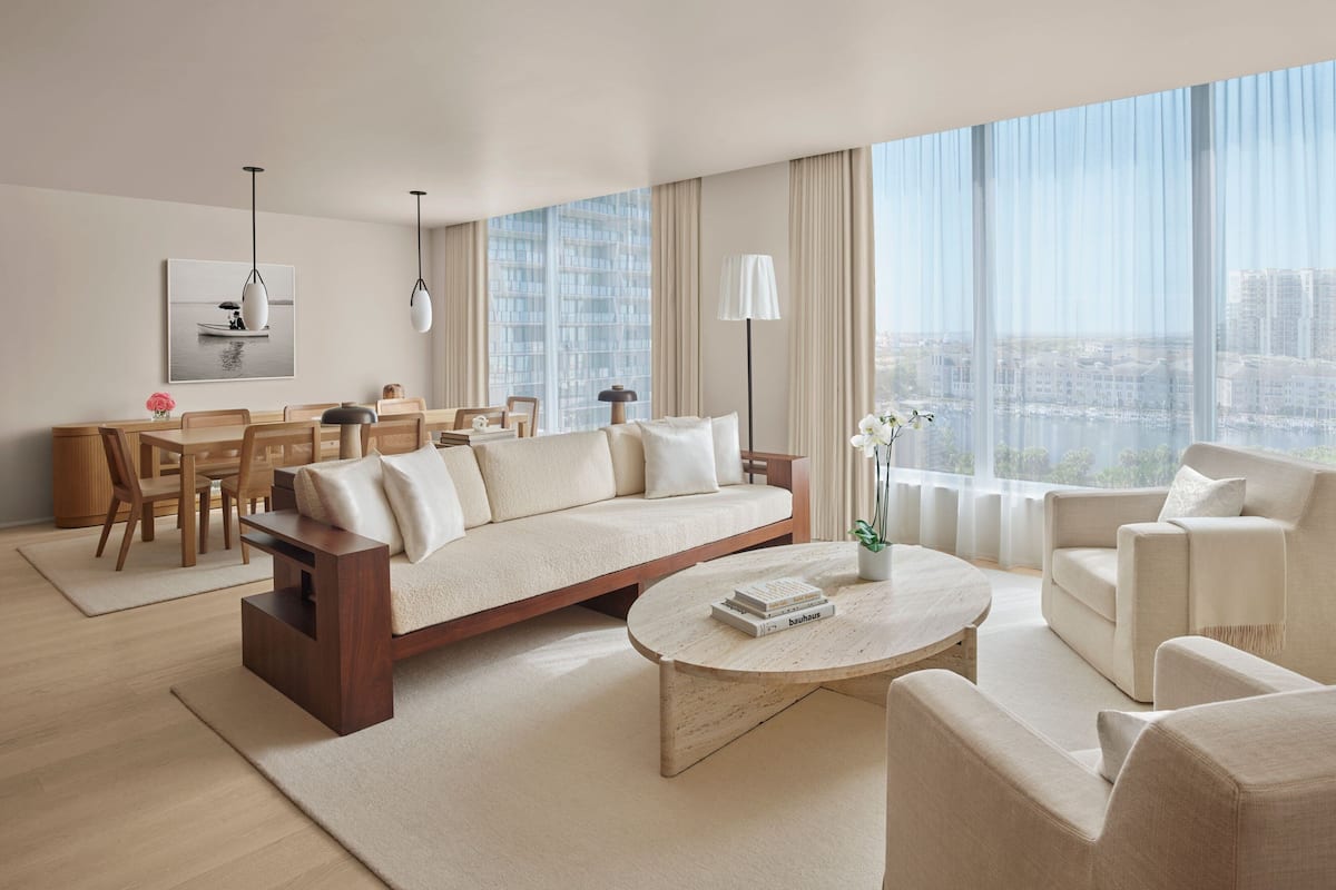 A modern living room with a sofa, coffee table, and chairs, a dining area with a table and chairs, and large windows offering a city view.