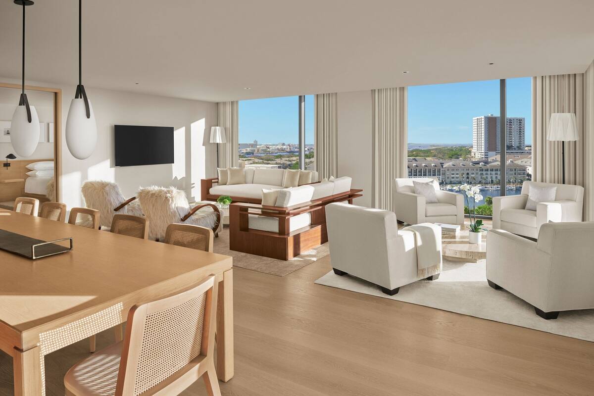 A modern hotel room with a large window offering a city view, a wooden dining table, and a comfortable seating area with sofas and chairs.