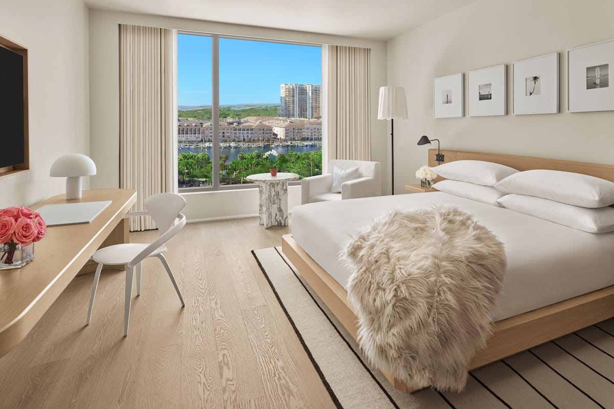 A modern hotel room with a large bed, a desk, a chair, and a view of the cityscape.
