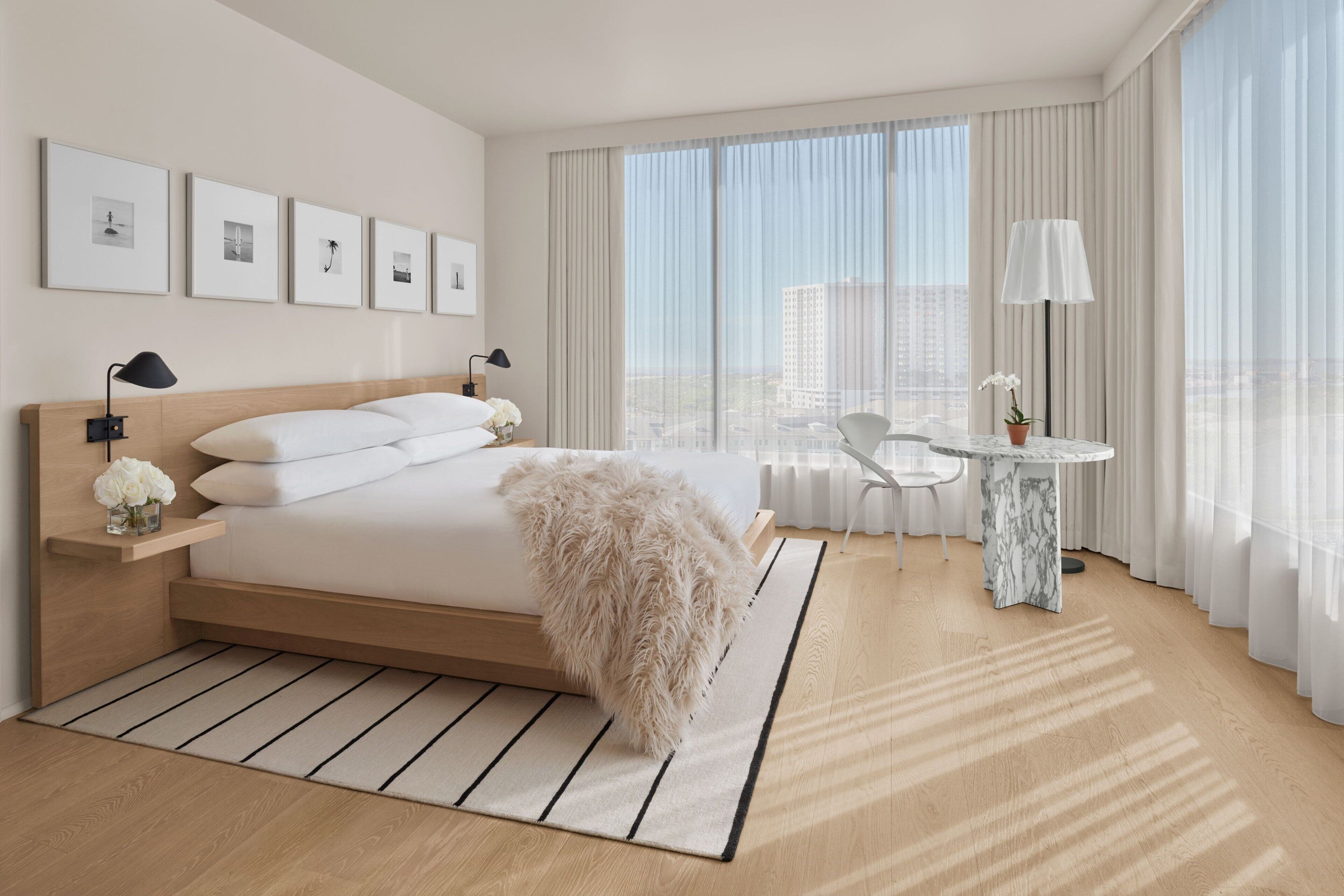 A modern hotel room with a large bed, a small table with a lamp, and a view of the city through large windows.