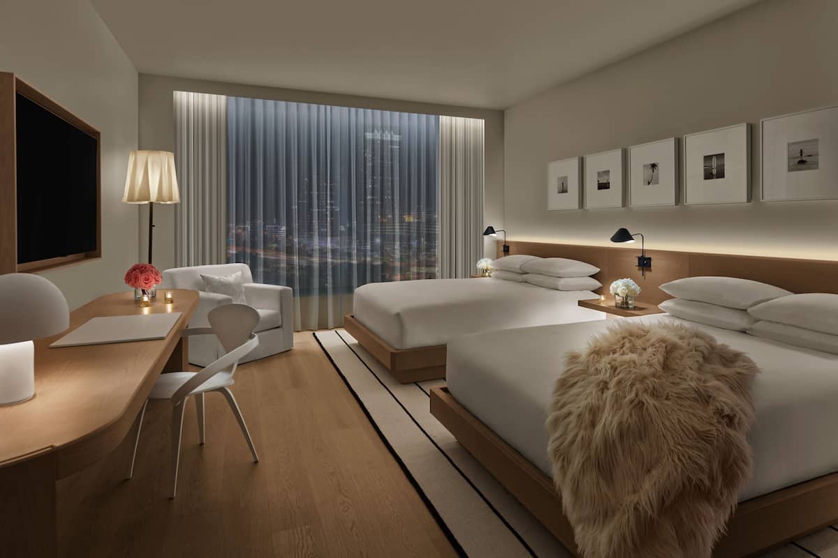 A modern hotel room with a large window, a desk with a chair, two beds, and a small table with a lamp.