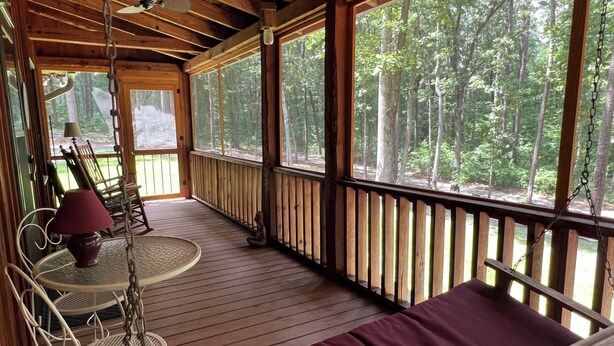 A wooden deck with a cushioned seating area, a table with chairs, and a view of a forest. - Cozy Cabin in the Woods, near Carolina Horse Park (Raeford)