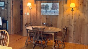A wooden-paneled room with a round table and four chairs, two wall-mounted lamps, and a painting of trees. - Cozy Cabin in the Woods, near Carolina Horse Park (Raeford)
