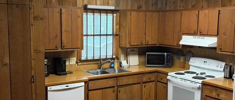 Una cocina con armarios de madera, un lavavajillas blanco, un microondas, una estufa y una ventana con persianas.