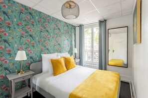 Habitación de hotel con cama, mesita de noche, lámpara, espejo y ventana con cortinas.