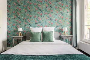 Un dormitorio con papel tapiz floral, dos mesitas de noche con lámparas y una cama con almohadas verdes y blancas.