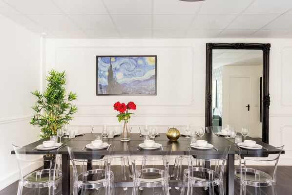 Un comedor con una larga mesa dispuesta para seis personas, sillas traslúcidas, un florero con flores rojas y un cuadro enmarcado sobre la mesa.