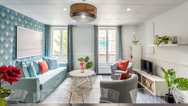 Een moderne woonkamer met een bank, stoelen, een salontafel en een televisie. Er staan planten en er is een raam met gordijnen.