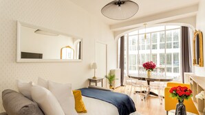 A hotel room with a bed, a sofa, a dining table with chairs, a vase with flowers, and a large window with curtains. - Merveil - Family Suite - Louvre - Sentier II (Paris)