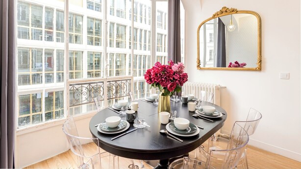 A dining room with a round table set for four, clear chairs, a vase of flowers, and a large mirror on the wall. - Merveil - Family Suite - Louvre - Sentier II (Paris)