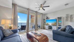 A modern living room with a large TV, two sofas, a wooden coffee table, and a balcony with a view of the beach.
