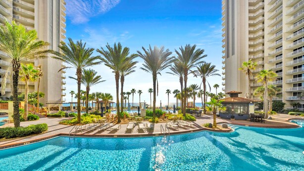 A resort pool area with palm trees, lounge chairs, and a view of the ocean.