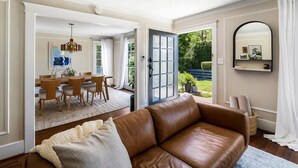A spacious living room with a brown leather sofa, a dining area with wooden chairs, and a large window offering a view of greenery.
