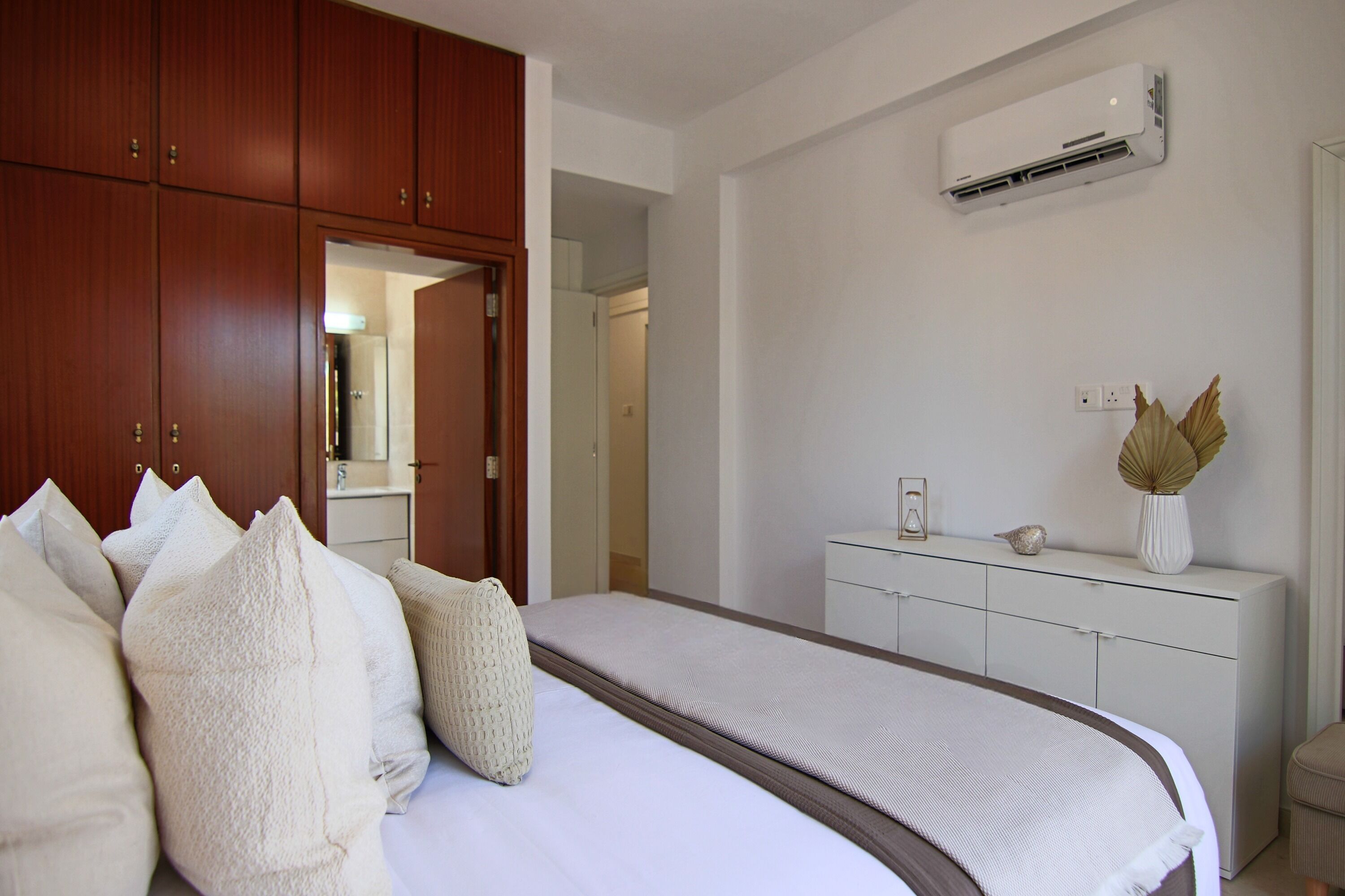 A bedroom with a bed, wooden wardrobe, white dresser, and an air conditioning unit.