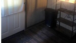 Una habitación con puerta de madera, cortina blanca, un bote de basura y una estantería con luz.