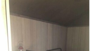 Una habitación pequeña y con poca luz, con una cama individual, una silla y una estantería con un objeto rojo.