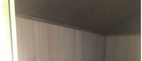Een kleine, zwak verlichte kamer met een enkel bed, een stoel en een plank met een rood voorwerp.
