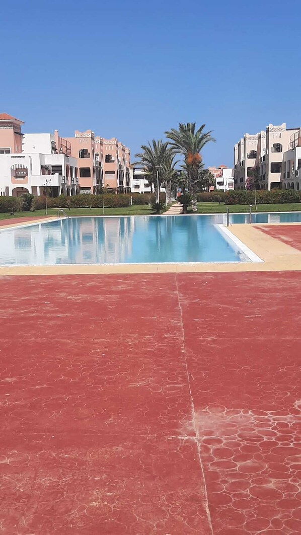 Piscina com piso vermelho, rodeada por edifícios com varandas e palmeiras.