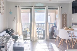 A living room with a sofa, a dining table with chairs, and a balcony with curtains. - Beachfront condo on the heart of Corpus Christi! (Corpus Christi)