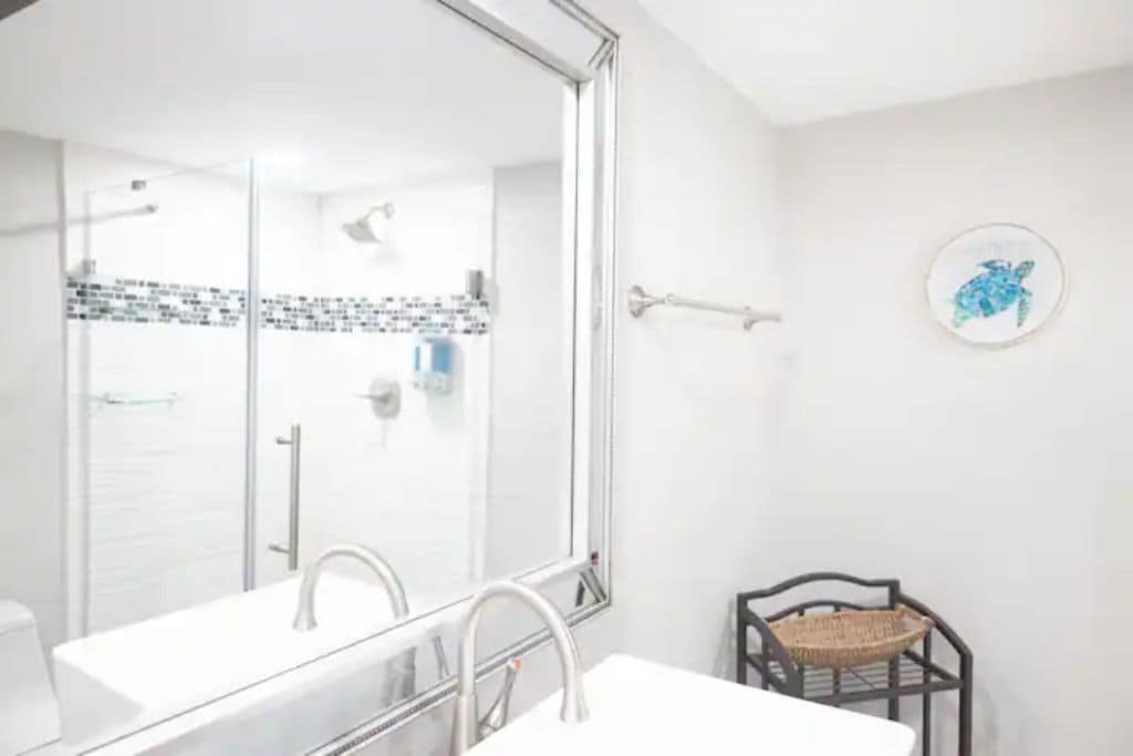 A bathroom with a large mirror, two sinks, a shower, and a small shelf with a basket.