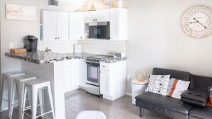 Eine moderne Küche mit weißen Schränken, einem eingebauten Backofen und einer Mikrowelle. Im angrenzenden Wohnbereich befinden sich weiße Hocker und eine schwarze Couch.