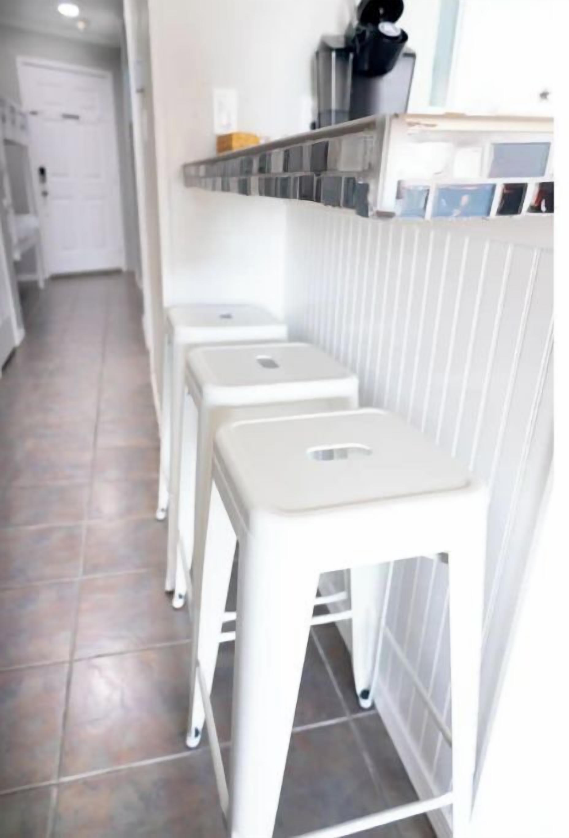 Eine Küche mit weiß gefliesten Wänden, einer weißen Arbeitsplatte und drei weißen Barhocker.