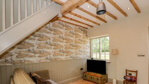A cozy living room with a high ceiling, exposed wooden beams, a stone-patterned wall, a large window, a television, a sofa, a chair, and a vintage suitcase. - The Hayloft at Warren House (Market Rasen)