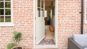 A brick house with a white door leading to an interior with a wooden floor and a wicker chair. - The Stable at Warren House (Market Rasen)