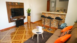 A living room with a TV, a coffee table, a sofa with orange cushions, a small table with a vase and remote controls, and a kitchen area in the background. - Ruinas: Spacious 2-bedroom apartment in San Blas market area. Big kitchen! (Cusco)