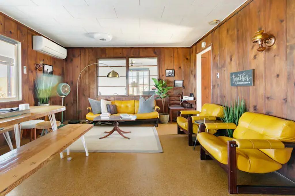 A cozy living room with wooden walls, yellow leather sofas, a wooden coffee table, and a dining area with a wooden table and chairs.