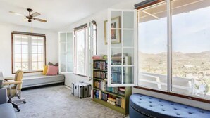 Una habitación con un ventanal grande, una estantería, un banco acolchado y un ventilador de techo.