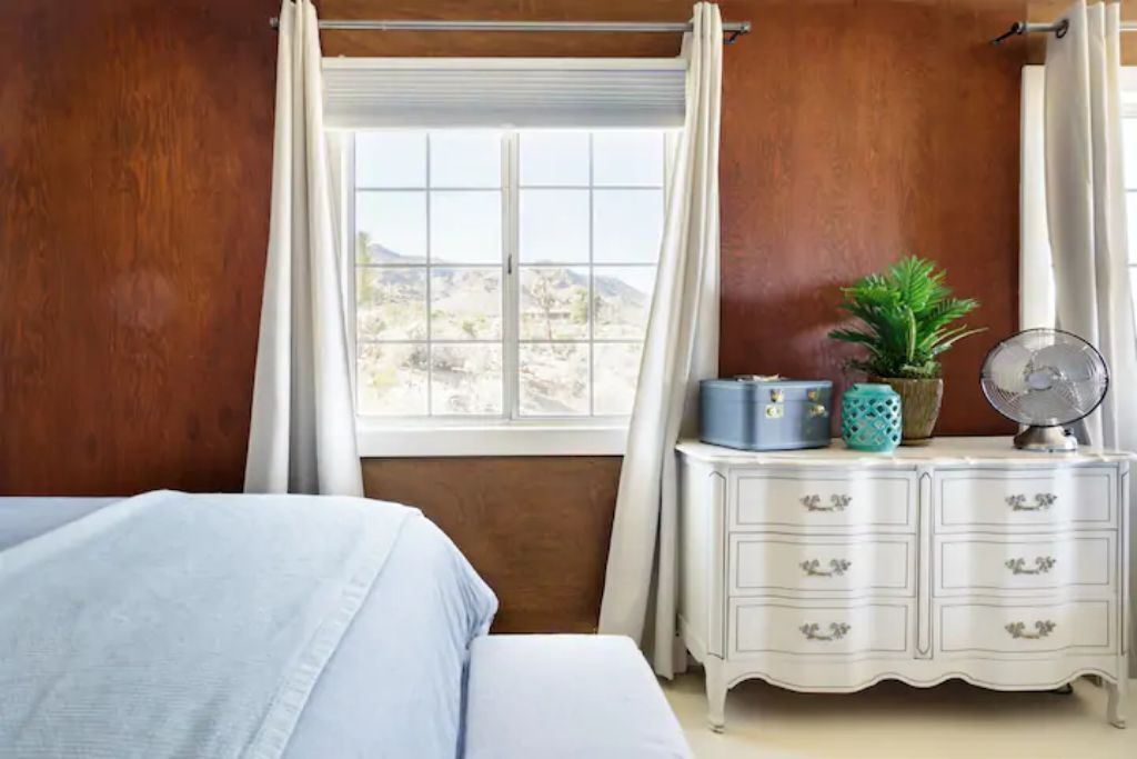 A bedroom with a bed, a dresser with drawers, a window with curtains, and a view of a desert landscape.