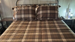 A bed with a plaid bedspread and two pillows, a metal headboard, and a small table with a lamp.
