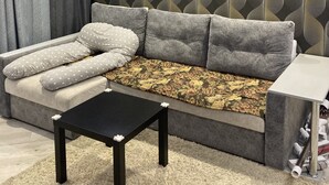 Uma sala de estar moderna com um sofá cinzento, almofadas estampadas, uma mesa de centro preta e uma parede decorativa com padrão de folha.