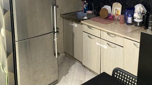 Uma cozinha compacta com geladeira de aço inoxidável, armários de madeira clara, bancada preta e um pequeno espaço de refeições com uma cadeira preta.