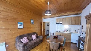 Een gezellige kamer met een houten plafond, een bank met gestreepte kussens, een eettafel met stoelen en een keuken met houten kastjes en een koelkast.
