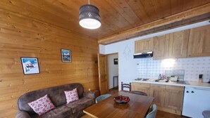 Een gezellige kamer met een houten plafond, een houten bank met gestreepte kussens, een houten eettafel met stoelen, en een keukenhoek met houten kastjes en een betegelde achterwand.